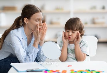 Cute little boy with speech problems practicing with professional therapist, doing practice and looking at mirror at cabinet, free space